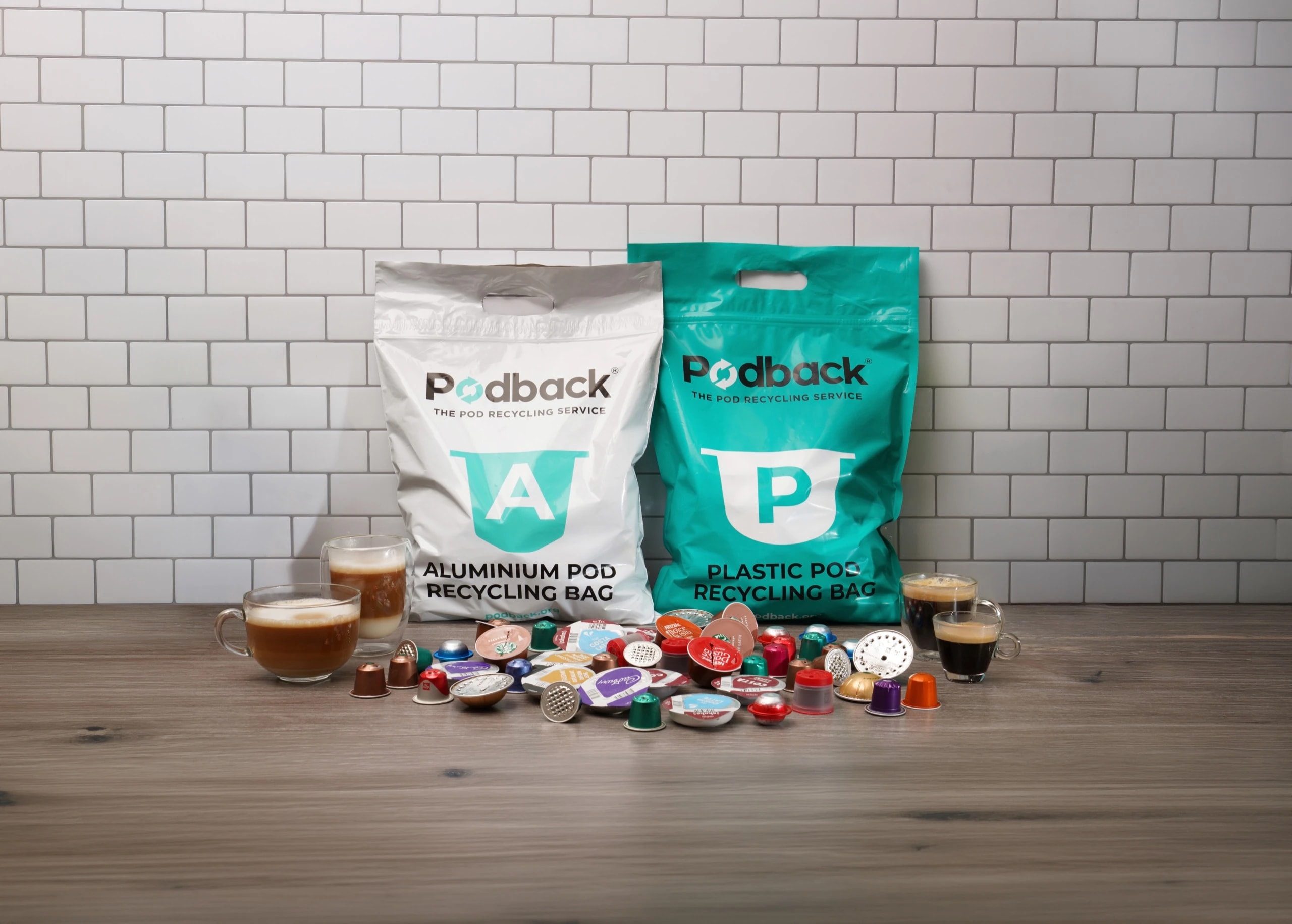 Podback aluminium and plastic pod recycling bags on a kitchen counter, surrounded by used coffee pods and cups of coffee