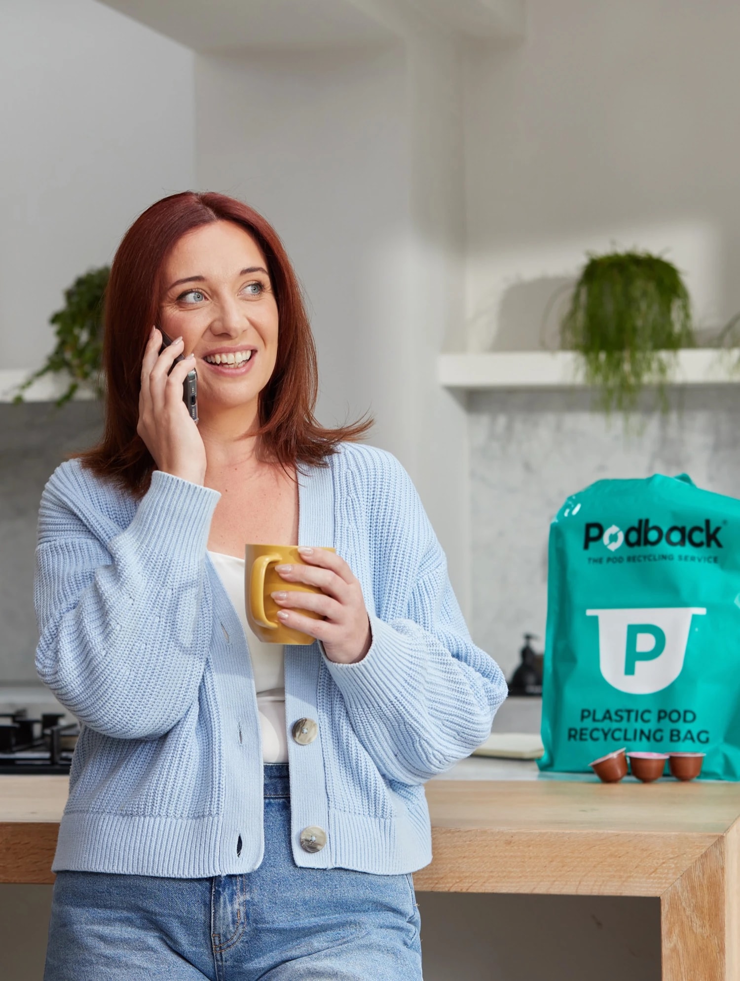 A woman enjoying a coffee in her kitchen beside a Podback plastic pod recycling bag