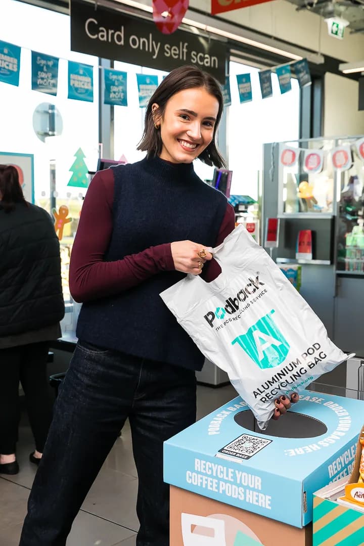 A smiling shopper posting a white Podback aluminium pod recycling bag into a Podback collection box in a supermarket